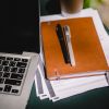 This image shows a laptop next to a pile of paper notes, notebook, and with a pair of pens on top