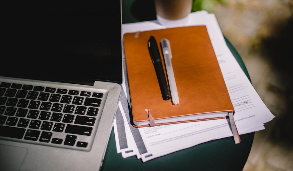 This image shows a laptop next to a pile of paper notes, notebook, and with a pair of pens on top