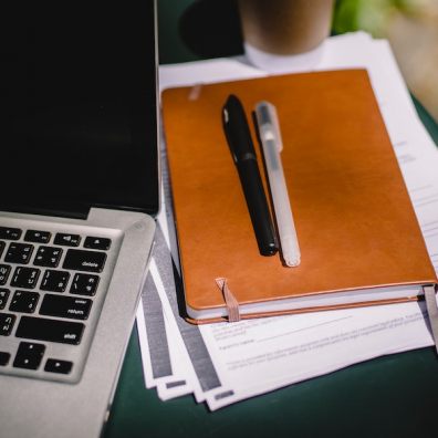 This image shows a laptop next to a pile of paper notes, notebook, and with a pair of pens on top